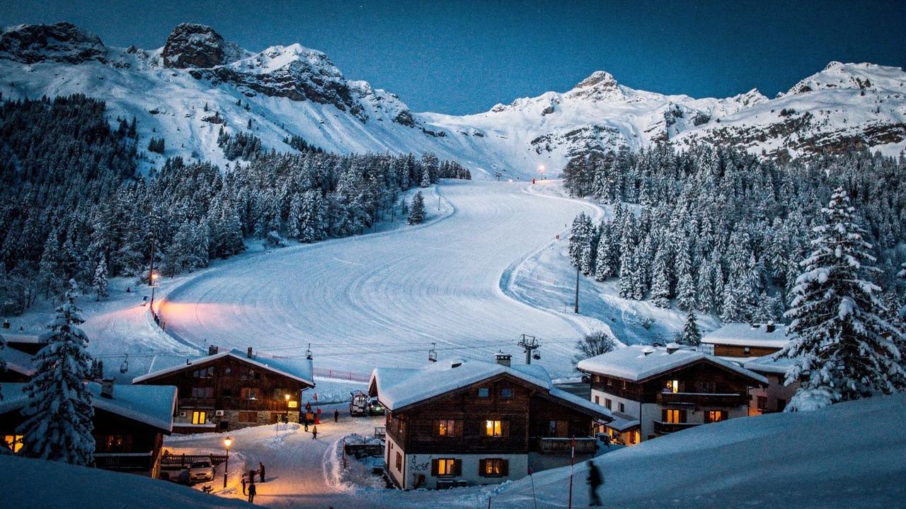 Alpine resort village at dusk with lit chalets and a groomed piste running down to the valley